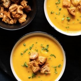 overhead shot of carrot soup served in two cream ceramic bowls on a black round board. topped with croutons and parsley with text layovers