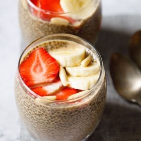 two glasses of chia pudding topped with sliced strawberries, bananas, pine nuts placed on a jute mat with two brass spoons by the side