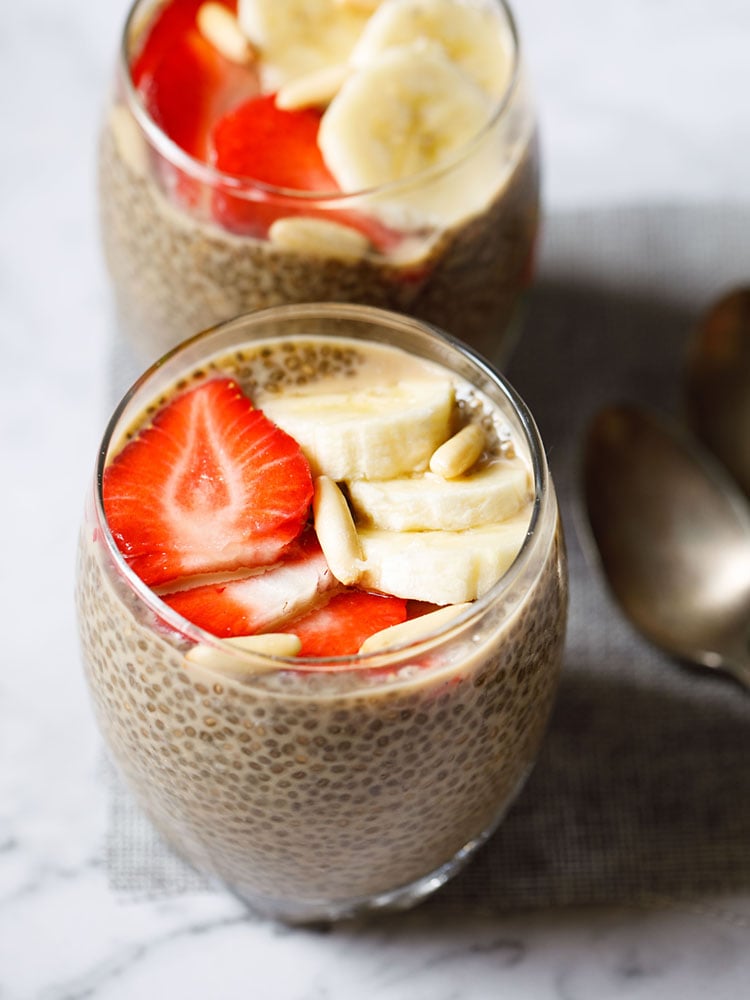 two glasses of chia pudding topped with sliced strawberries, bananas, pine nuts placed on a jute mat with two brass spoons by the side.