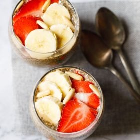 top shot of two glasses of chia pudding topped with sliced strawberries, bananas, pine nuts placed on a jute mat with two brass spoons by the side