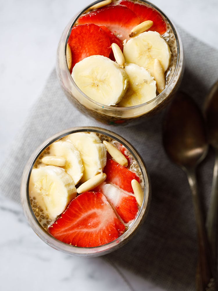 topped with sliced bananas, strawberries, pine nuts the two glasses of chia seed pudding are placed on a gray jute mat on a white marbled board.