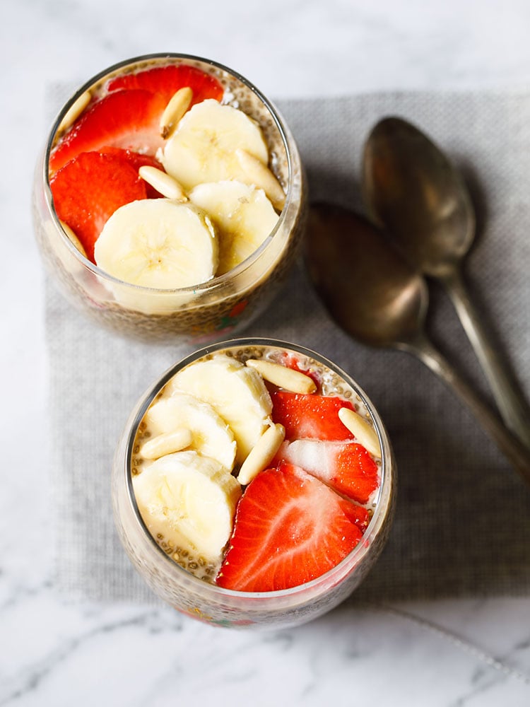 Top shot of two glasses of chia pudding, topped with sliced ​​strawberries, bananas, pine nuts, on a jute mat with two brass spoons on the side.