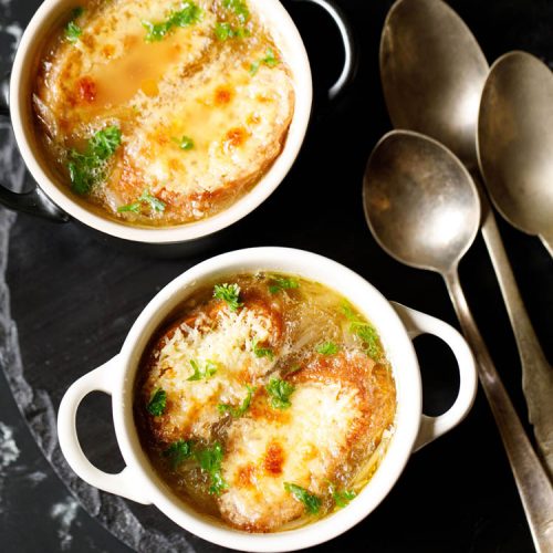 french onion soup in two cream colored soup bowls with spoons on a black slate tray. soup garnished with some parsley.