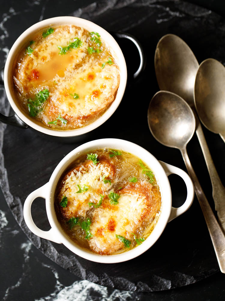 french onion soup in two cream colored soup bowls with spoons on a black slate tray. soup garnished with some parsley.