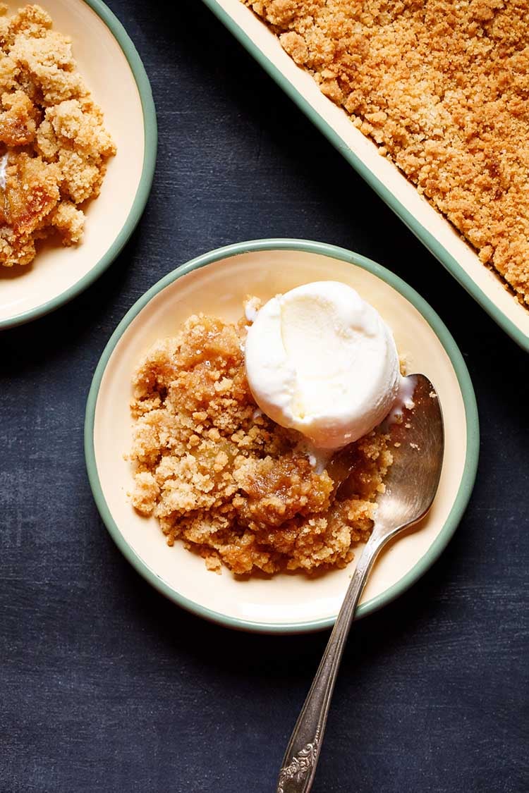apple crumble topped with a scoop of vanilla icecream in a green rimmed cream plate with a dessert spoon