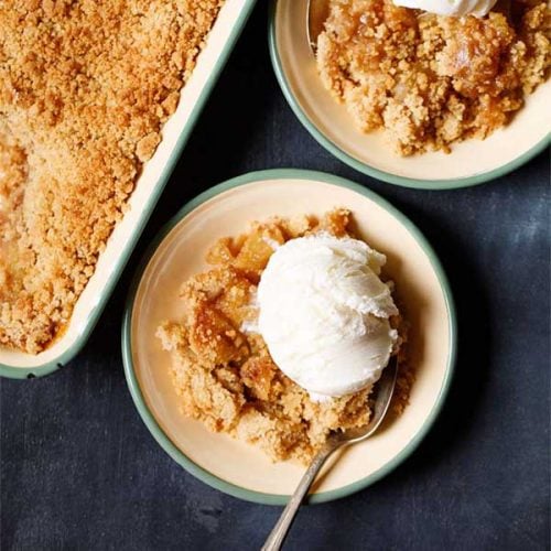 apple crumble topped with a scoop of vanilla icecream in a green rimmed cream plate with a dessert spoon.
