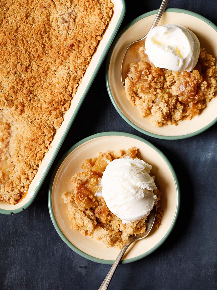 apple crumble topped with a scoop of vanilla icecream in a green rimmed cream plate with a dessert spoon