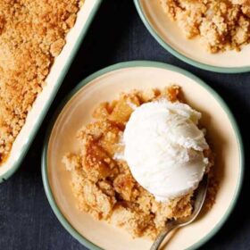 apple crumble topped with a scoop of vanilla icecream in a green rimmed cream plate with a dessert spoon.