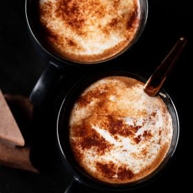 homemade hot chocolate sprinkled with cocoa powder served in 2 black cups (with a copper spoon in one cup) on a black marble board