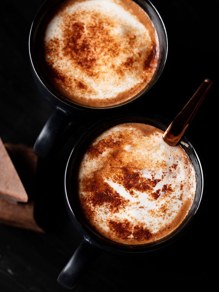 homemade hot chocolate sprinkled with cocoa powder served in 2 black cups (with a copper spoon in one cup) on a black marble board