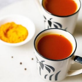 Turmeric Tea served in off white small cups with a black flower design on the cup. Cups placed on a white marble board with a small bowl or turmeric and a few black pepper thrown on the marble board
