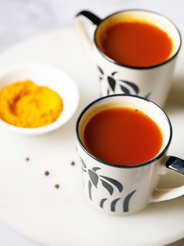 Turmeric Tea served in off white small cups with a black flower design on the cup. Cups placed on a white marble board with a small bowl or turmeric and a few black pepper thrown on the marble board