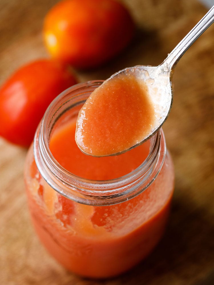 tomato puree in a spoon on top of a glass jar filled with tomato puree and two tomatoes placed on top left side