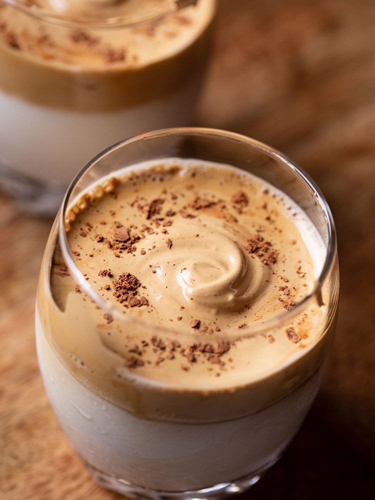 a glass of whipped coffee sprinkled with cocoa powder and placed on a brown wooden board. 