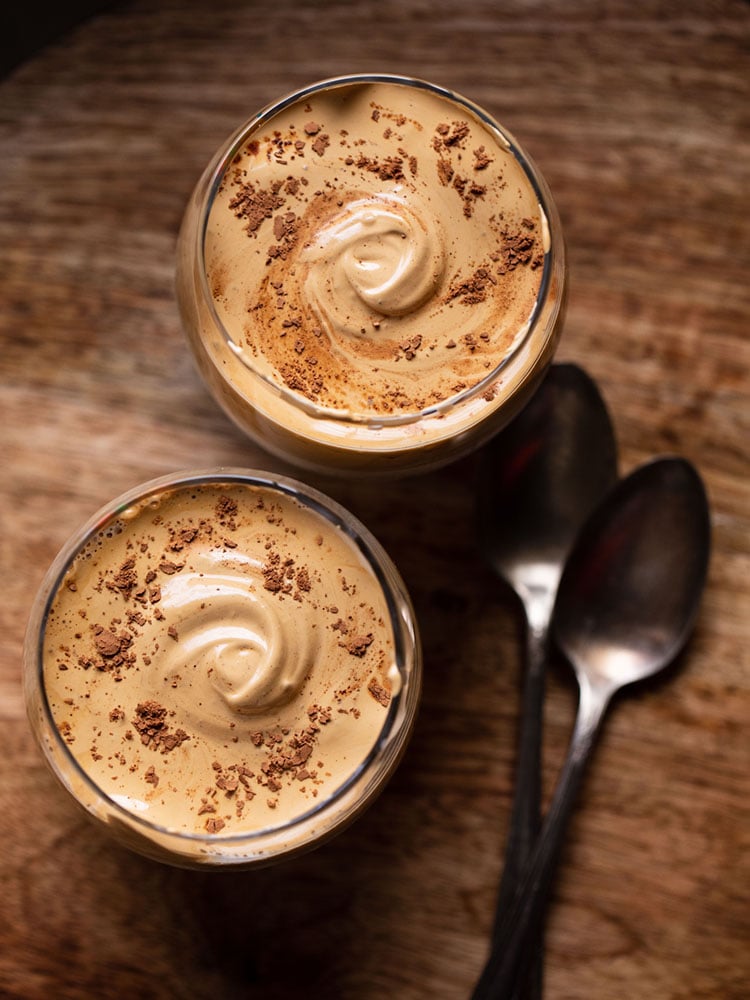 dalgona coffee topped with some cocoa powder and served in two glasses on a brown wooden board. 