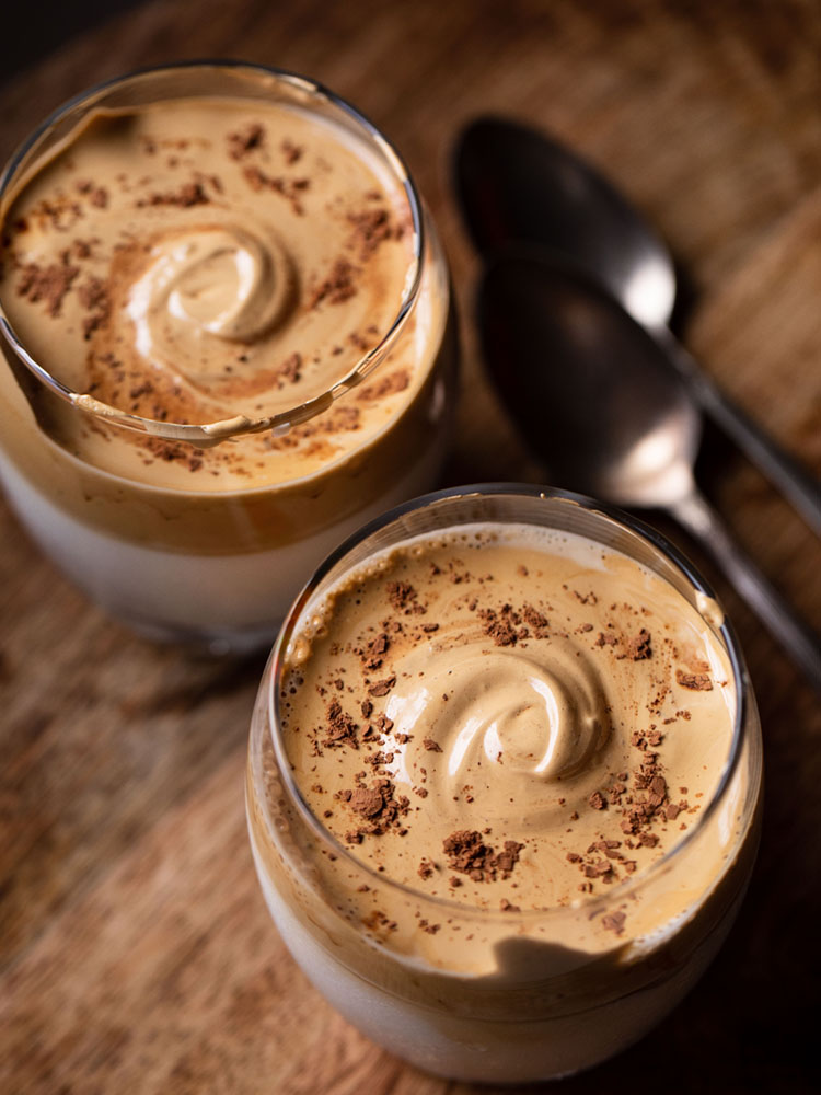 whipped coffee topped with some cocoa powder and served in two glasses on a brown wooden board