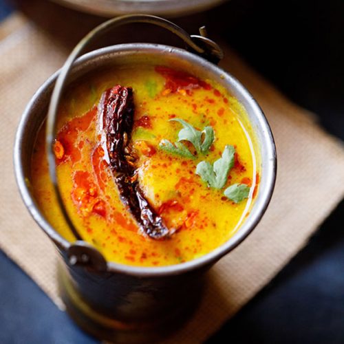 dal tadka garnished with cilantro and a topped with fried red chilli with some fried cumin and some red colored oil in a small brass bucket on a light brown jute mat