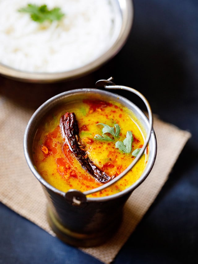 dal tadka garnished with cilantro and a topped with fried red chilli with some fried cumin and some red colored oil in a small brass bucket on a light brown jute mat with a bowl of steamed rice kept above on the top-right.