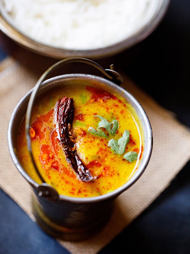 dal tadka garnished with cilantro and a topped with fried red chilli with some fried cumin and some red colored oil in a small brass bucket on a light brown jute mat with a bowl of steamed rice kept above on the top-right.