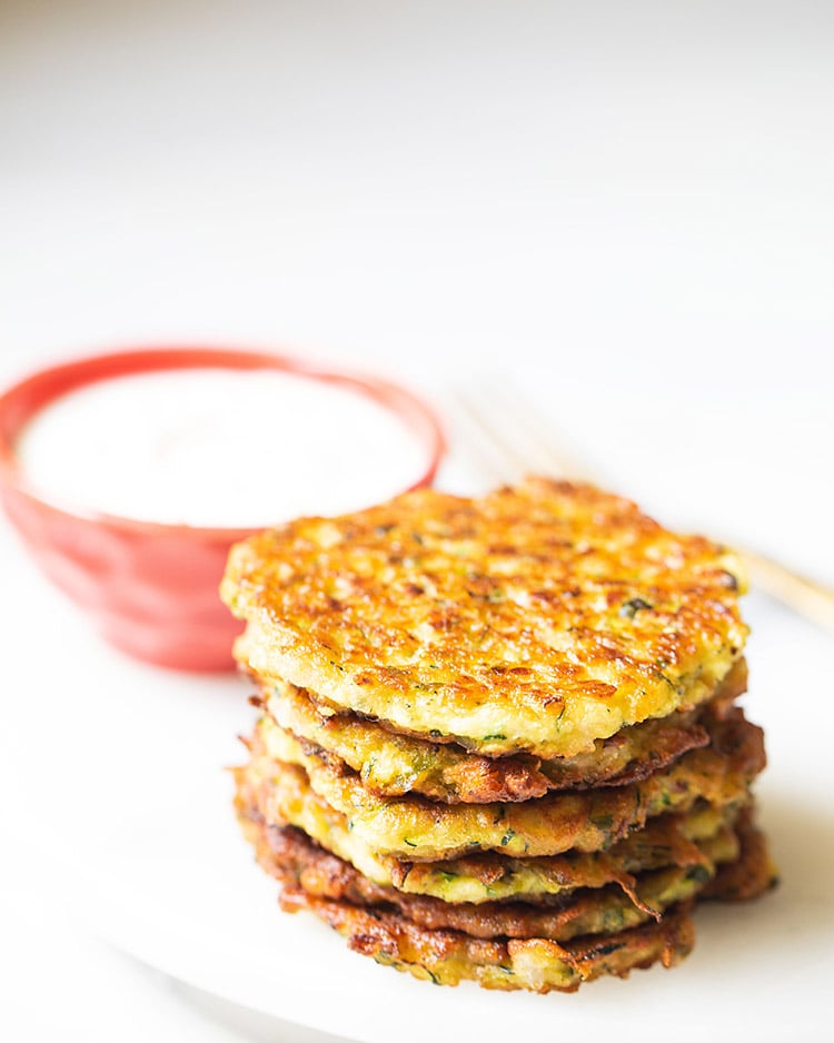 zucchini fritters stacked and kept on white marble board with sour cream in a red bowl kept in the background.