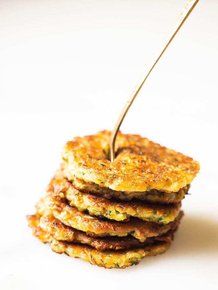 zucchini fritters stacked on top of each other with a brass fork inside them placed on a white marble board.