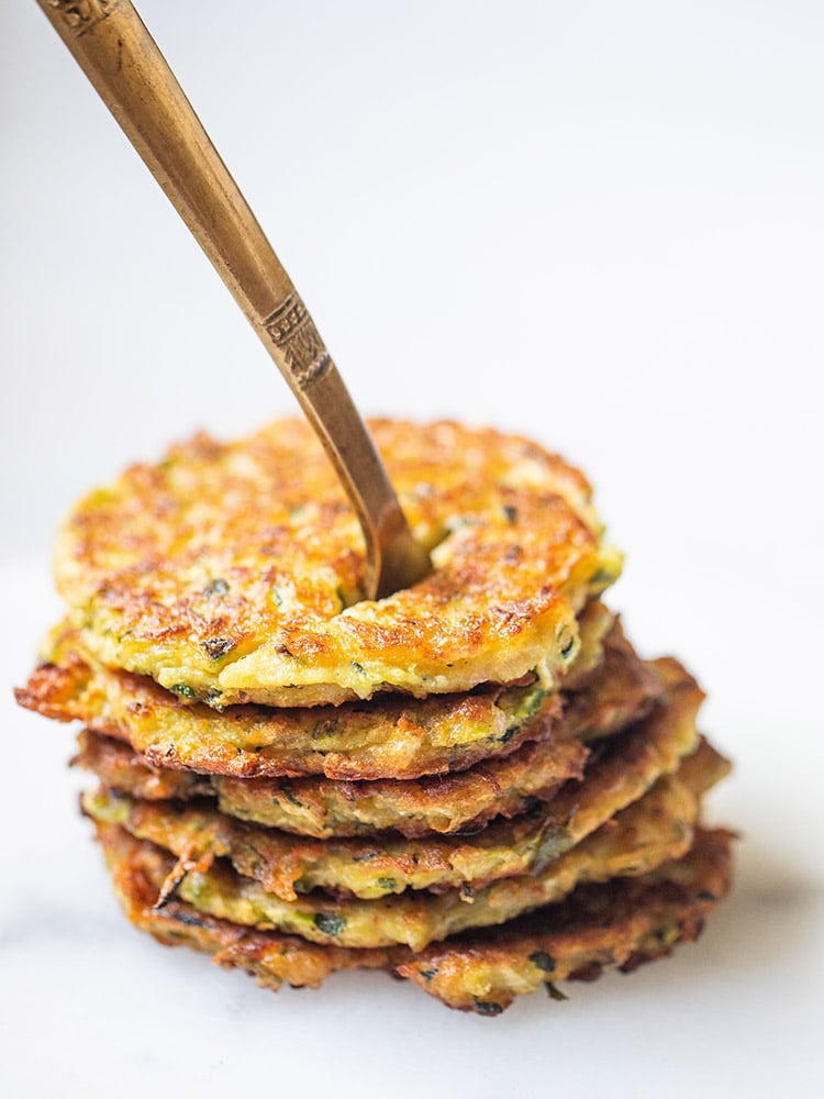 zucchini fritters stacked on top of each other with a brass fork inside them placed on a white marble board