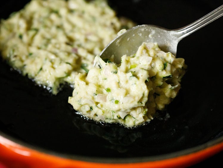 portion of zucchini fritter batter in a spoon on top of skillet containing hot oil.