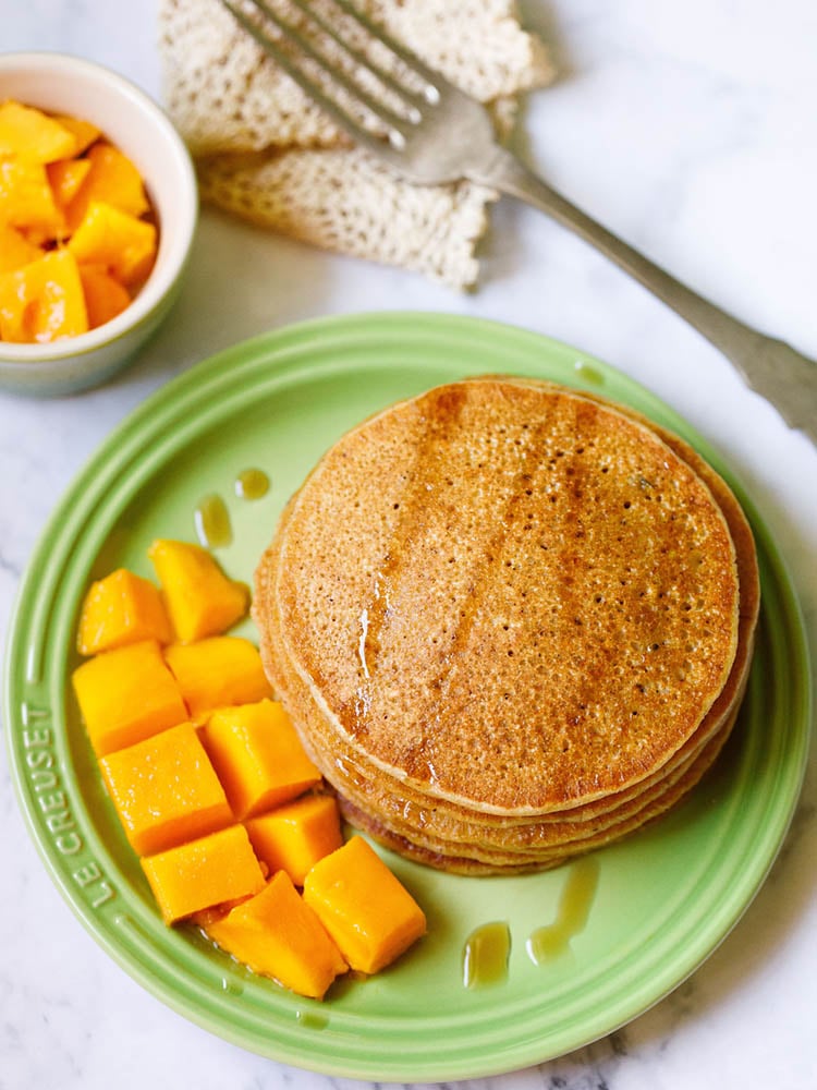 pumpkin pancakes stacked on green plate drizzled with maple syrup with a side of cubed mangoes, a small bowl of mangoes on top right and with a fork kept on folded crochet doily near top left on a white marble background.