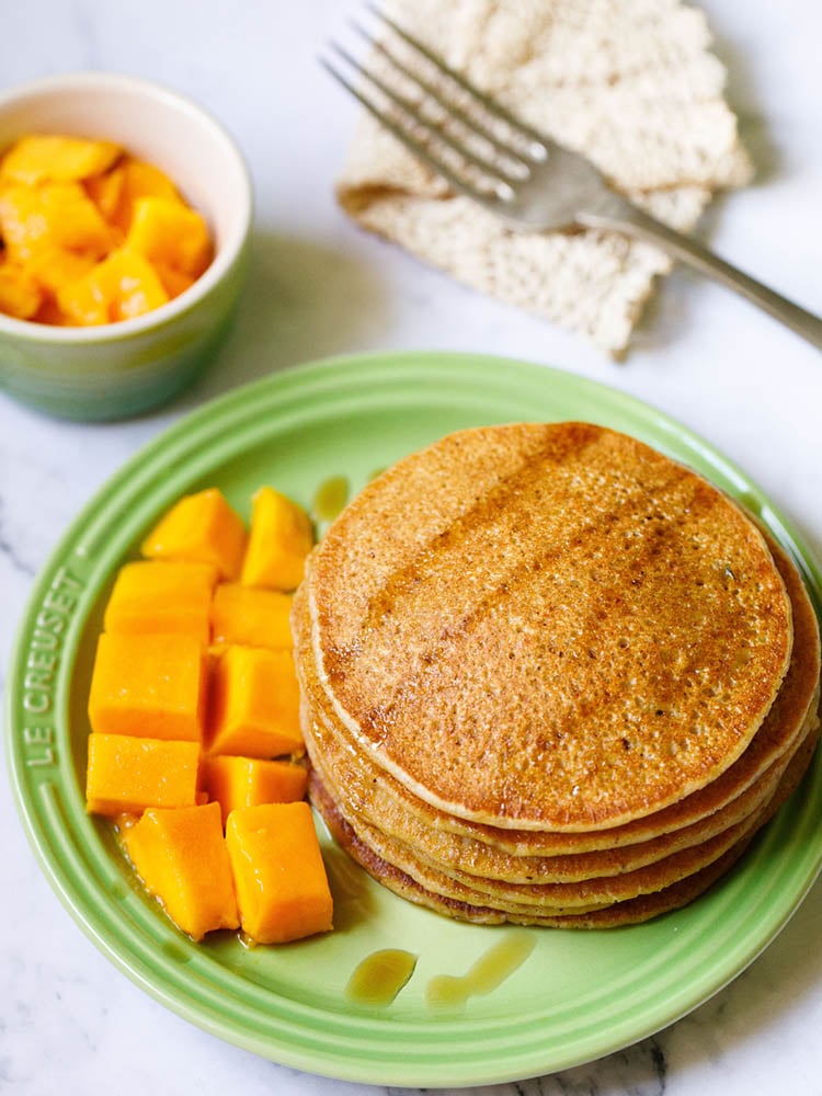 pumpkin pancakes stacked on green plate drizzled with maple syrup with a side of cubed mangoes and a small bowl of cubes mangoes on the top.