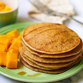 pumpkin pancakes stacked on green plate drizzled with maple syrup with a side of cubed mangoes