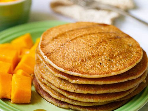 pumpkin pancakes stacked on green plate drizzled with maple syrup with a side of cubed mangoes