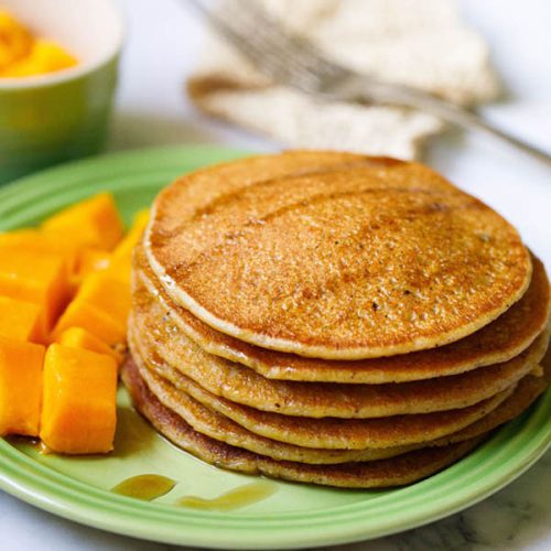 pumpkin pancakes stacked on green plate drizzled with maple syrup with a side of cubed mangoes