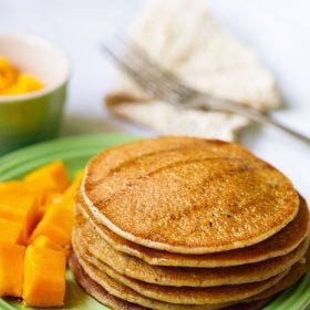 pumpkin pancakes stacked on green plate drizzled with maple syrup with a side of cubed mangoes