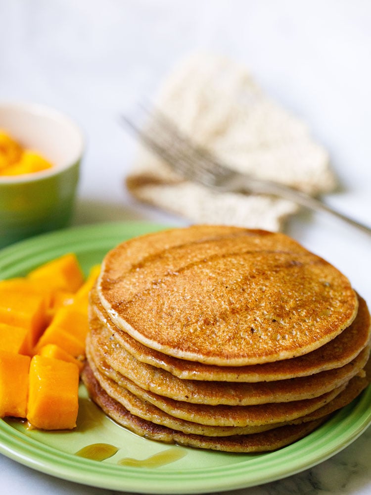 pumpkin pancakes stacked on green plate drizzled with maple syrup with a side of cubed mangoes.