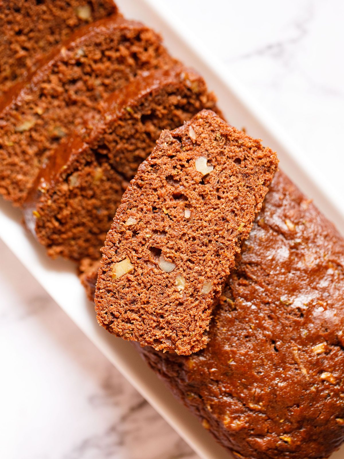 chocolate zucchini bread partly sliced with one slice kept on top of the loaf on a white rectangular tray on a white marble background