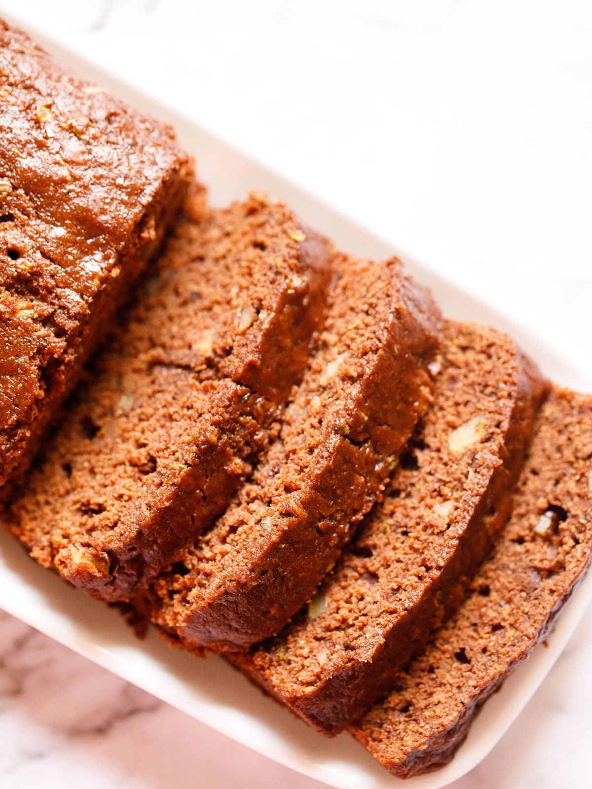 chocolate zucchini bread sliced in four slices and the entire loaf and the slices are kept on a white rectangular tray on a white marble background