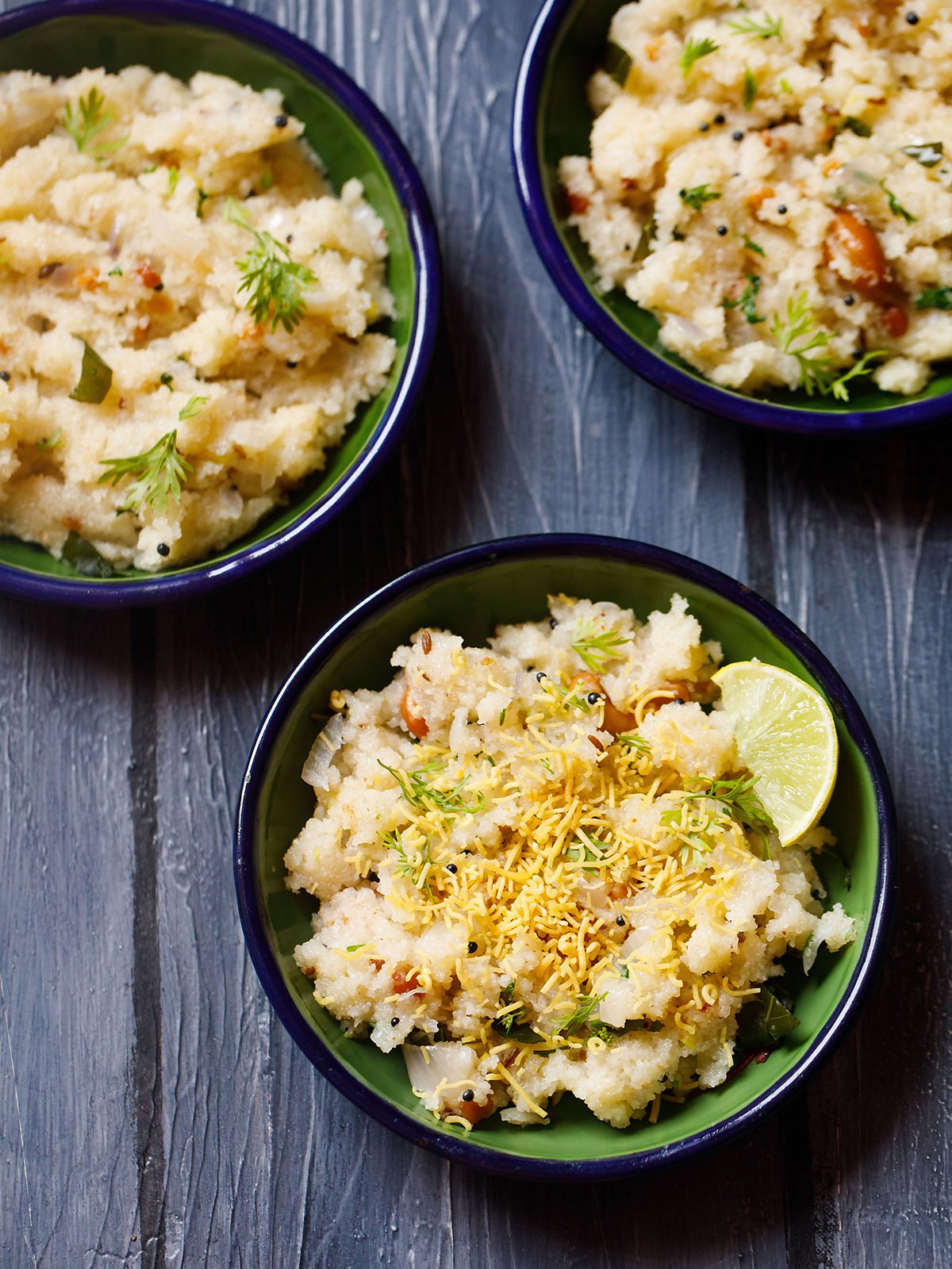 rava upma garnished with cilantro, fried gram flour vermicelli and a lemon wedge served in a dark green blue rimmed plate on a dark blue wooden board