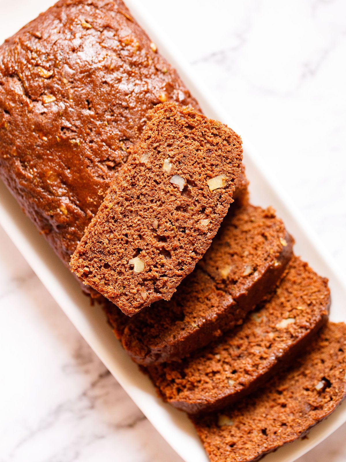 zucchini bread partly sliced with one slice kept on top of the loaf on a white rectangular tray on a white marble background