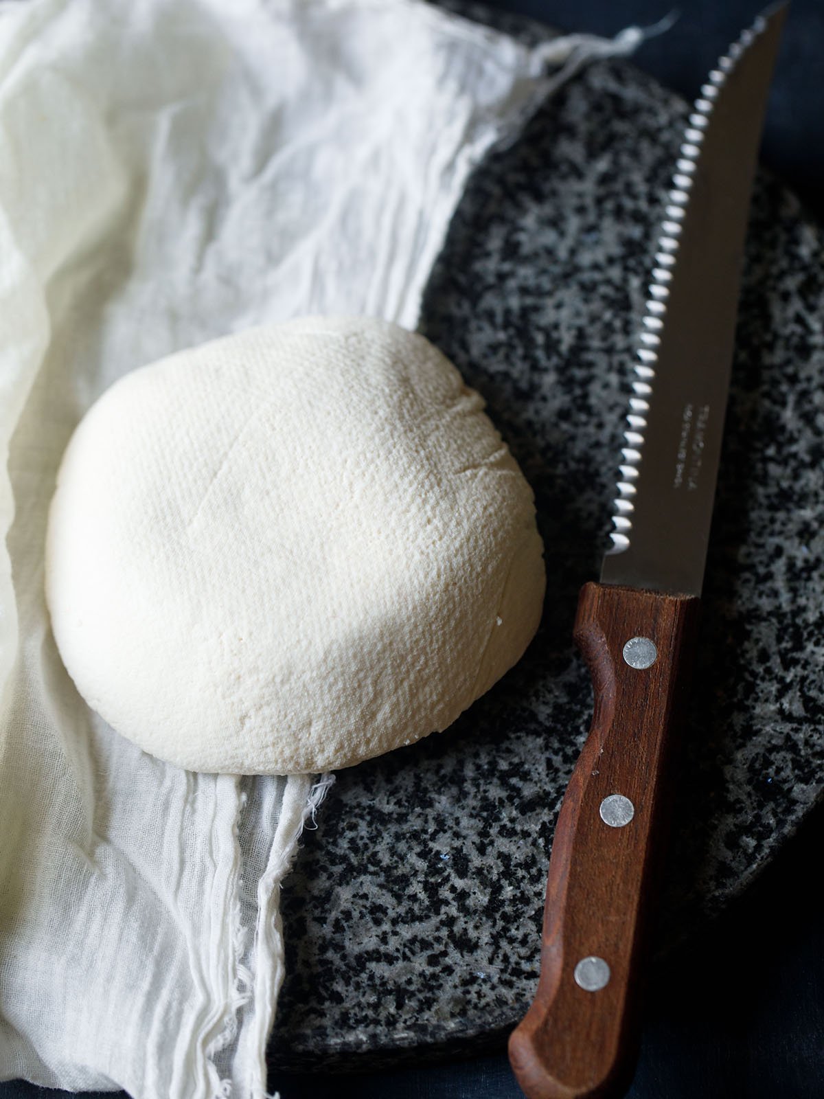 round block of homemade paneer on a white muslin with a serrated wooden knife at side on a black-grey granite board 