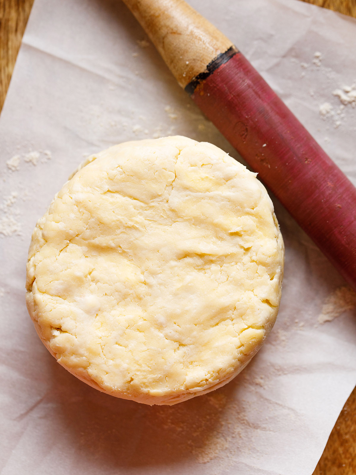 round of all butter pie dough on a floured piece of parchment with a red rolling pin on the side prior to rolling out the dough