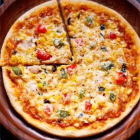Whole round Pizza on a wooden pizza plate with a cut triangular slice on left top side