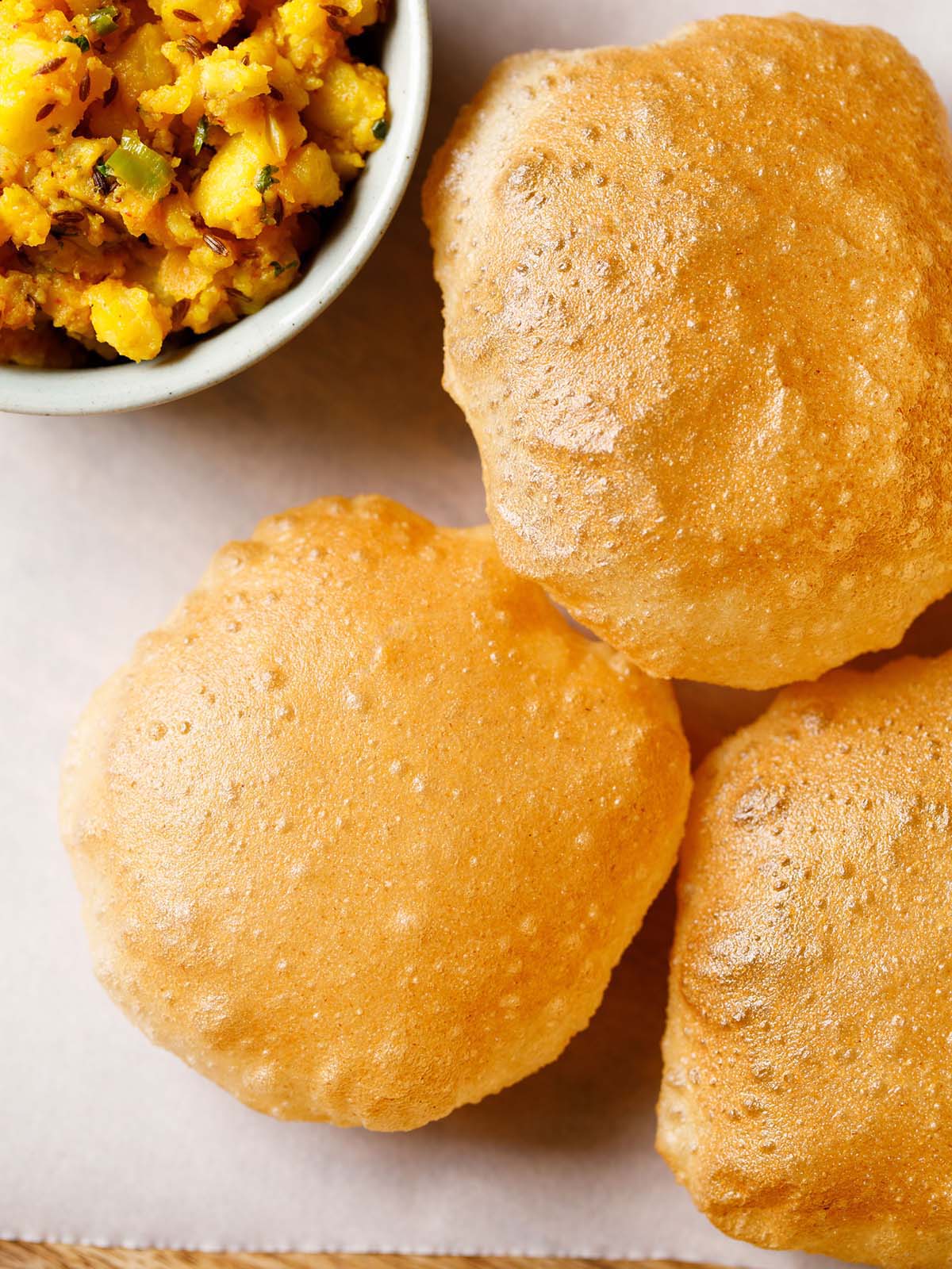 homemade puri placed on parchment paper with a side bowl of spiced sautéed Indian potato dish