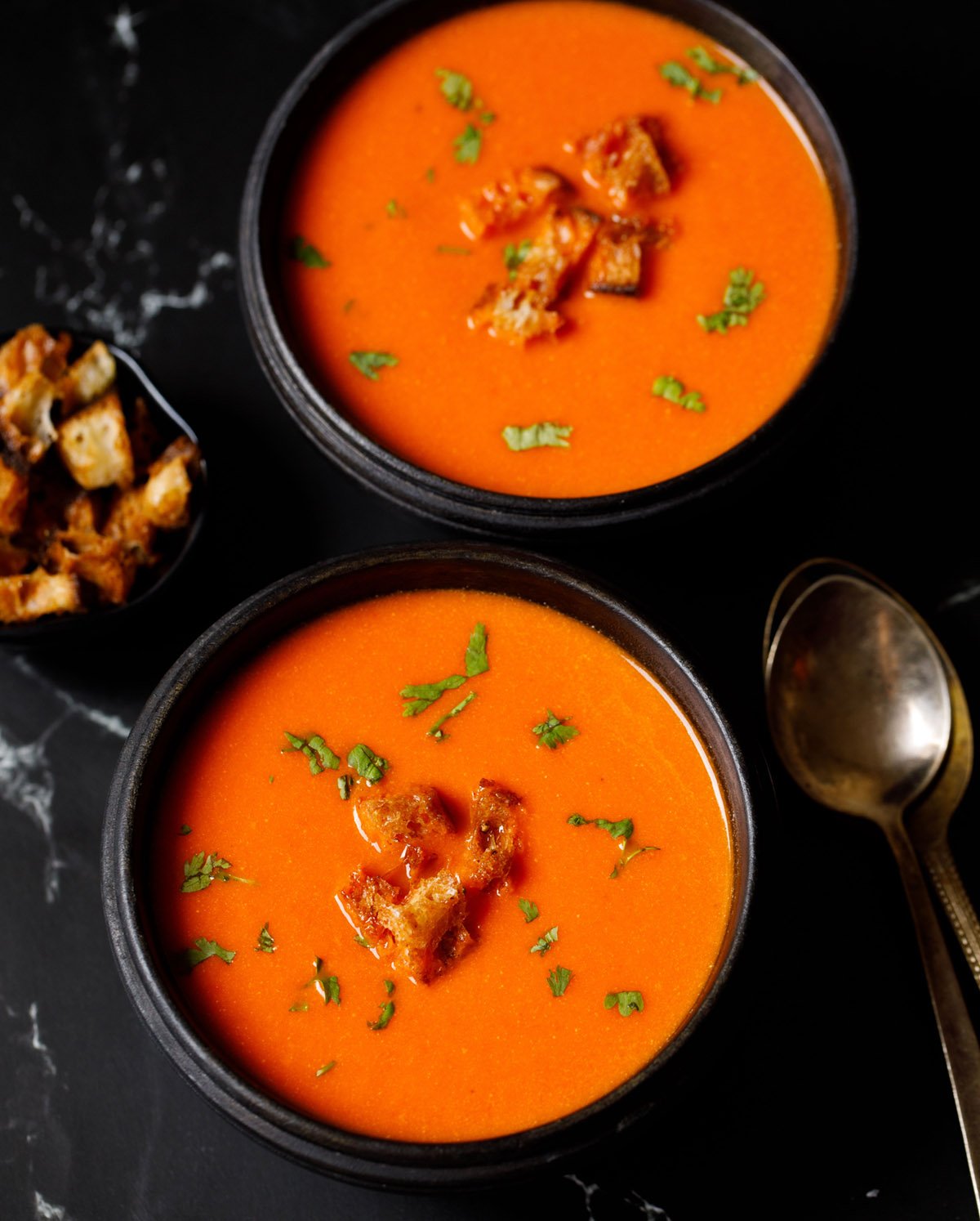 tomato soup topped with croutons, parsley in two black wooden bowls with silver and brass spoons by the side on a black marble