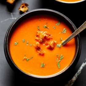 tomato soup in a black wooden bowl with a silver spoon and topped with bread croutons and parsley