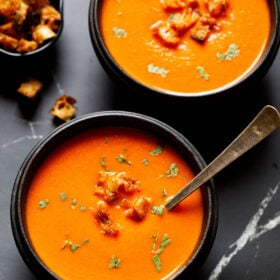 tomato soup in a black wooden bowl with a silver spoon topped with croutons and parsley