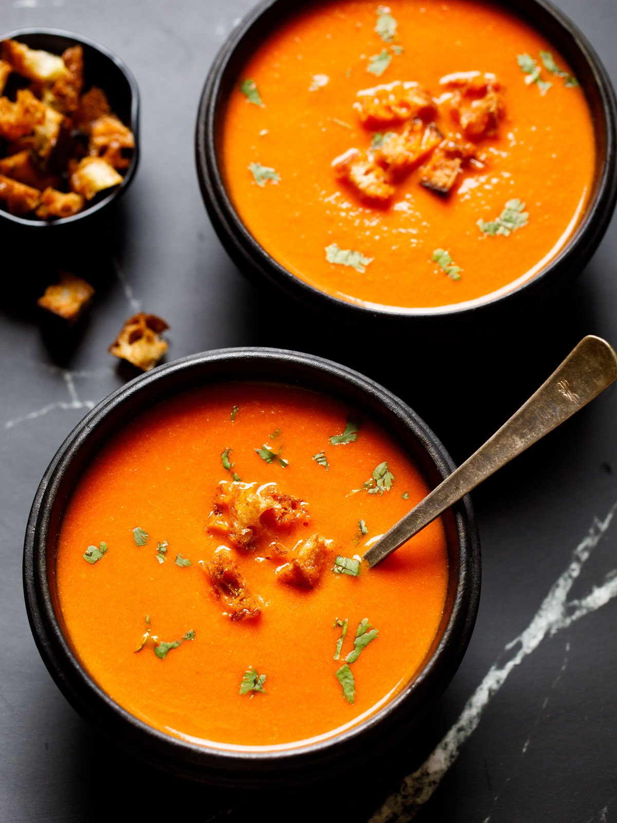 tomato soup in a black wooden bowl with a silver spoon topped with croutons and parsley