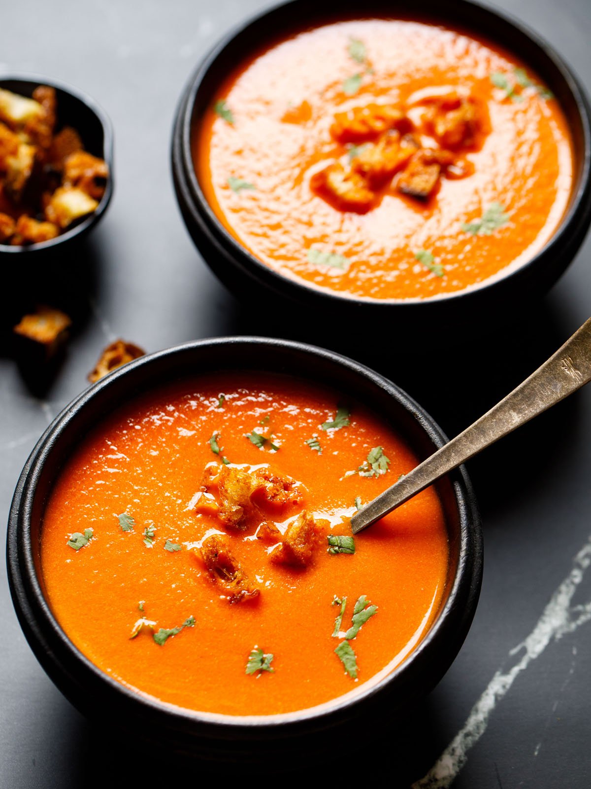 homemade rustic tomato soup from scratch in a black wooden bowl with a silver spoon topped with croutons and parsley