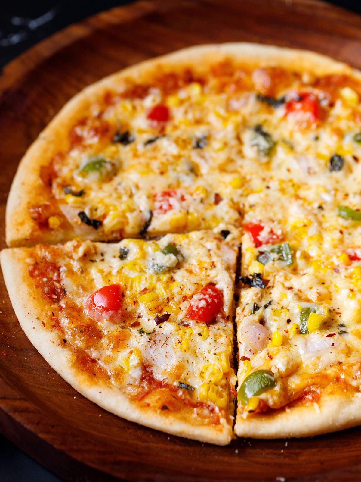 a triangular slice of veg pizza with the remaining pizza on a wooden pizza plate sprinkled with some dried herbs and red chilli flakes