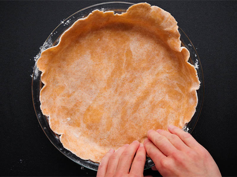 rolled pie dough placed in a glass pie pan and edges being pushed into the pan