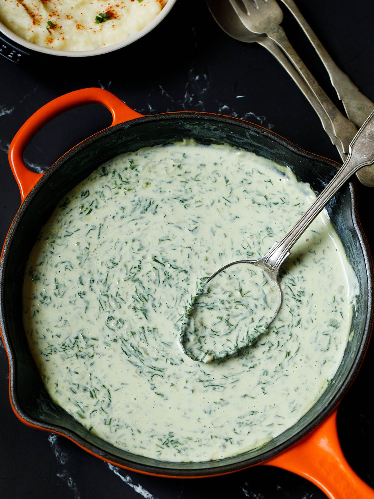 overhead shot of finished creamed spinach recipe in an orange/red enameled cast iron skillet with a silver serving spoon